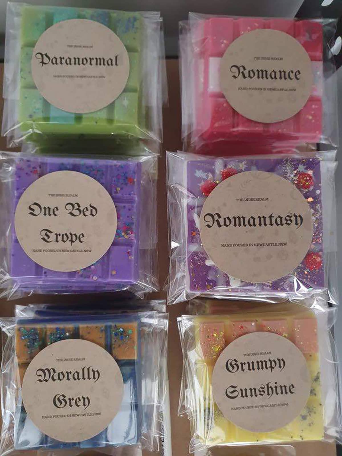 Six wax melt blocks; "Paranormal" wax melt in green, "Romance" wax melt in pink, "One Bed Trope" wax melt in lilac, "Romantasy" wax melt in purple, "Morally Grey" wax melt in orange and grey, "Grumpy Sunshine" wax melt in yellow and pink