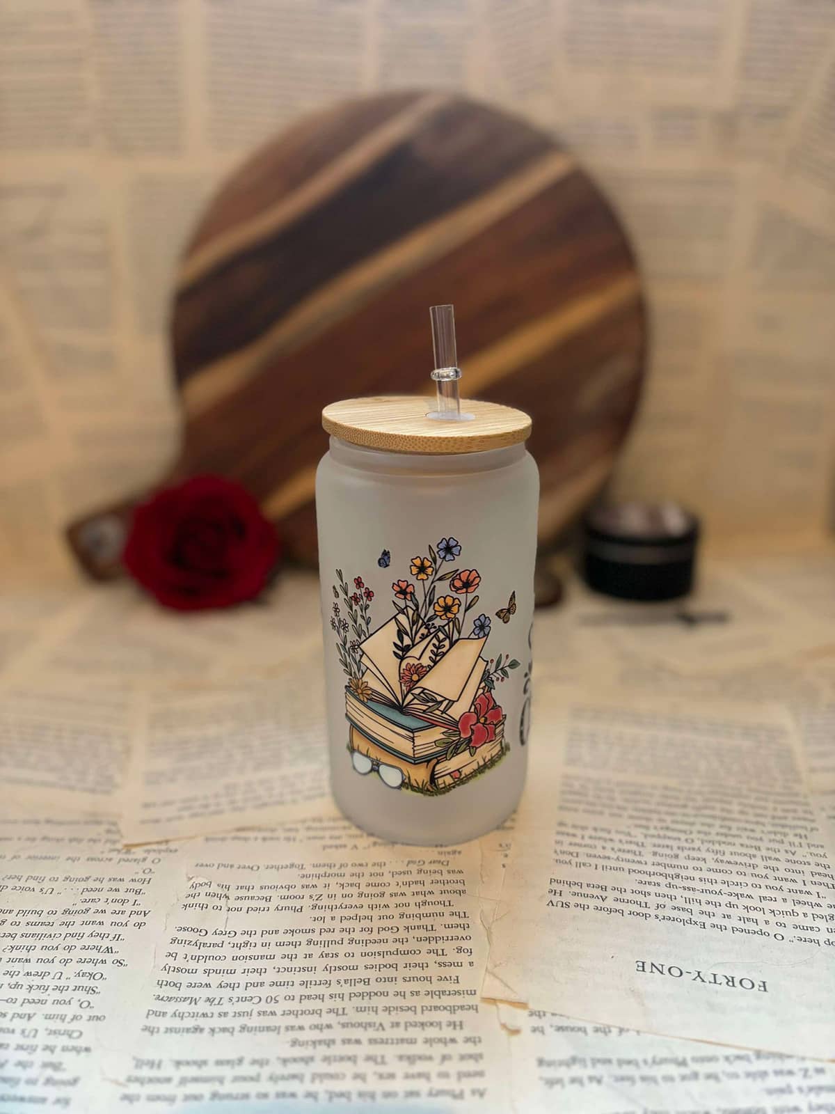 Just One More Chapter themed frosted glass cup featuring illustration of colourful stack of books with flowers growing through on frosted glass, with wooden lid and clear straw