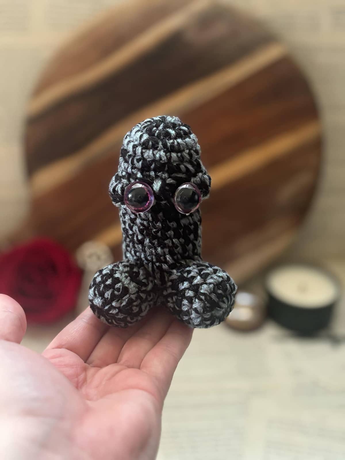 Hand holding crochet pin cushion in penis shape, featuring purple eyes on blue, grey and black yarn