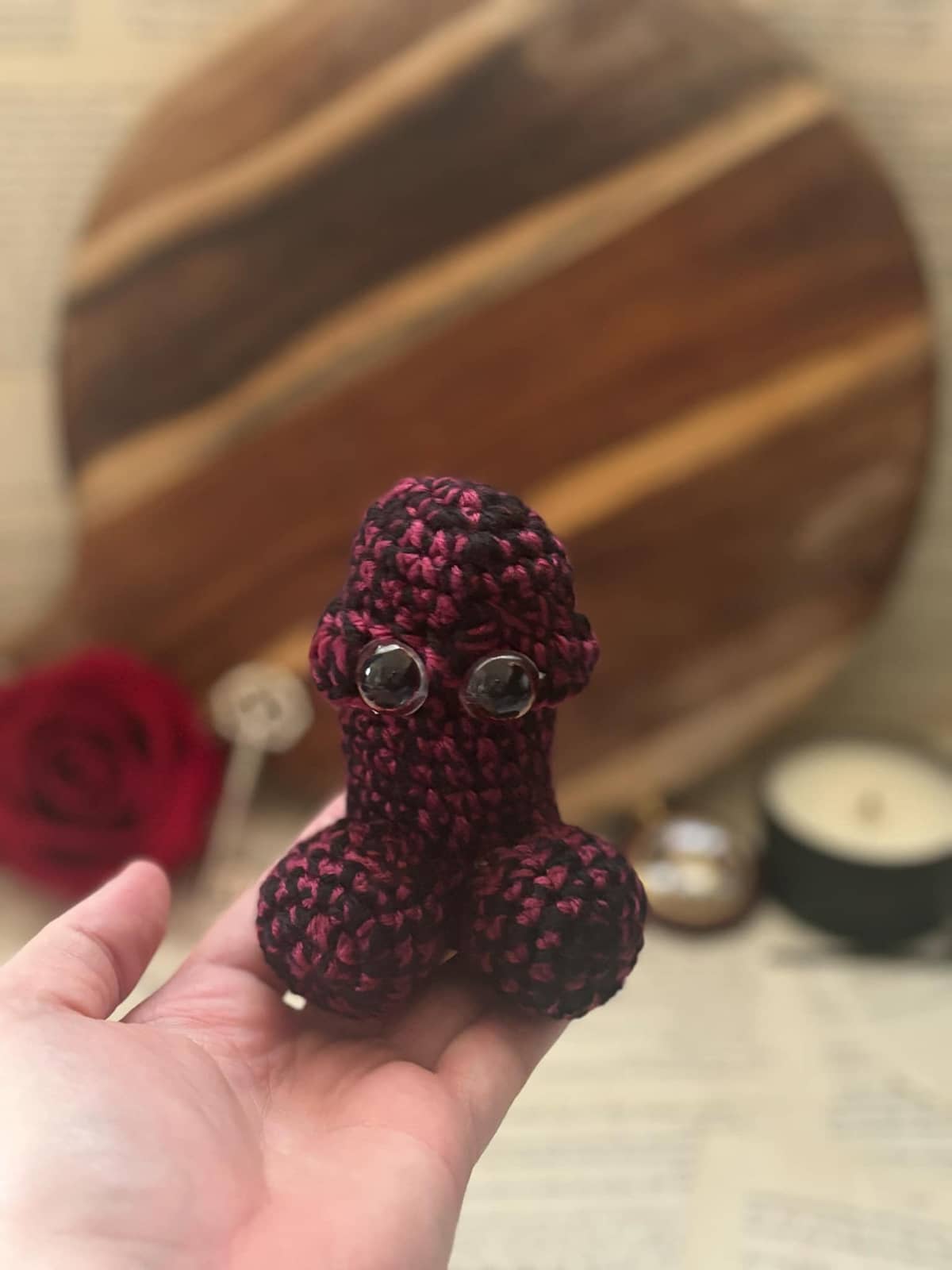 Hand holding crochet pin cushion in penis shape, featuring eyes on dark red and black yarn