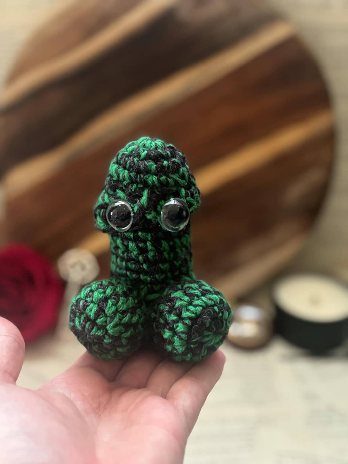 Hand holding crochet pin cushion in penis shape, featuring eyes on green and black yarn