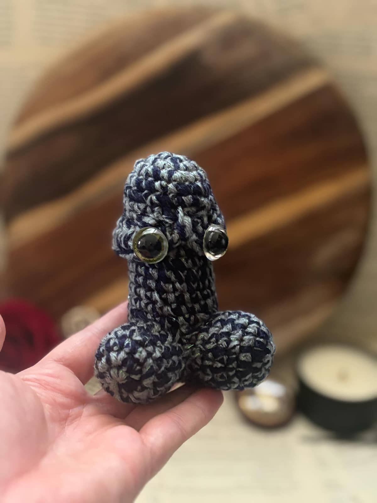 Hand holding crochet pin cushion in penis shape, featuring eyes on grey and navy yarn