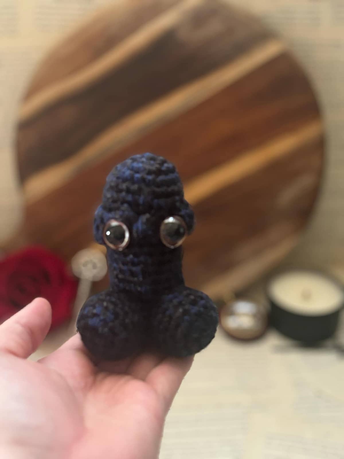 Hand holding crochet pin cushion in penis shape, featuring eyes on navy and black yarn