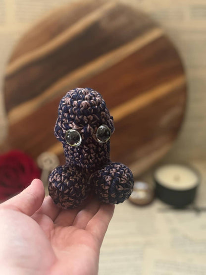 Hand holding crochet pin cushion in penis shape, featuring eyes on pink and navy yarn