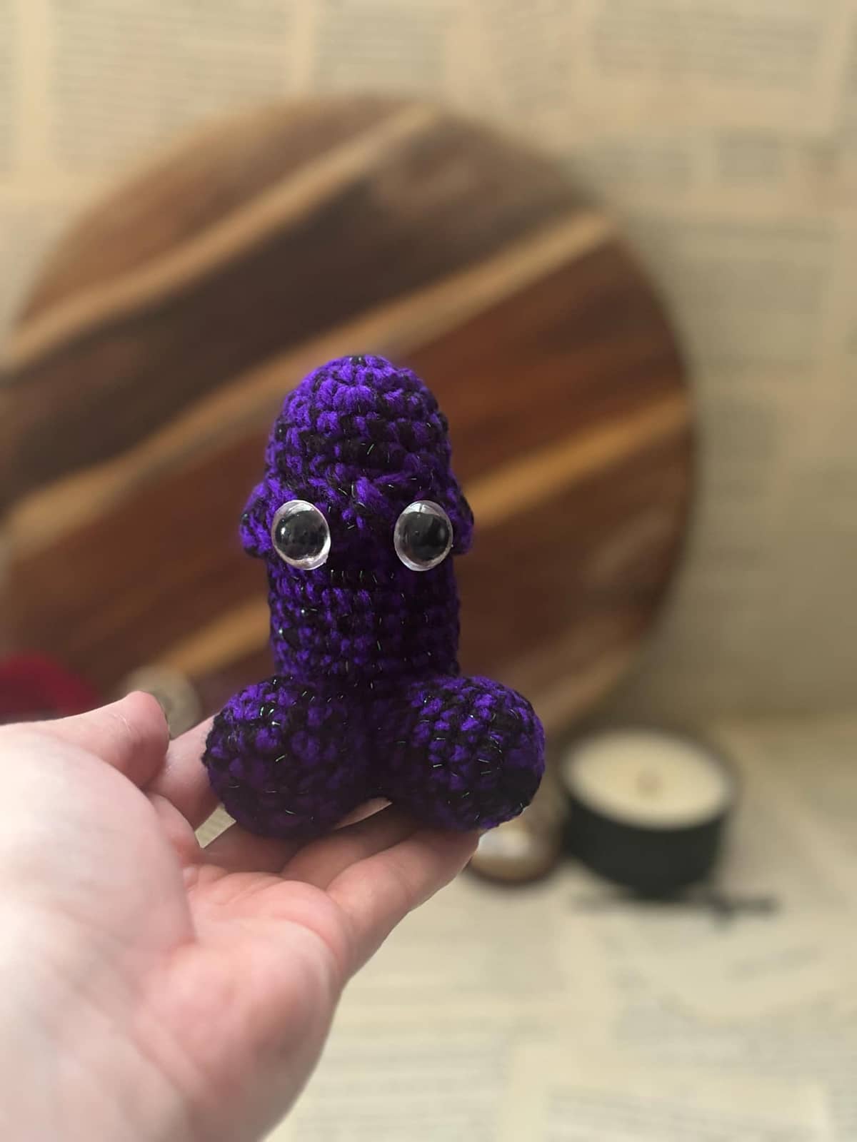 Hand holding crochet pin cushion in penis shape, featuring eyes on purple and black yarn