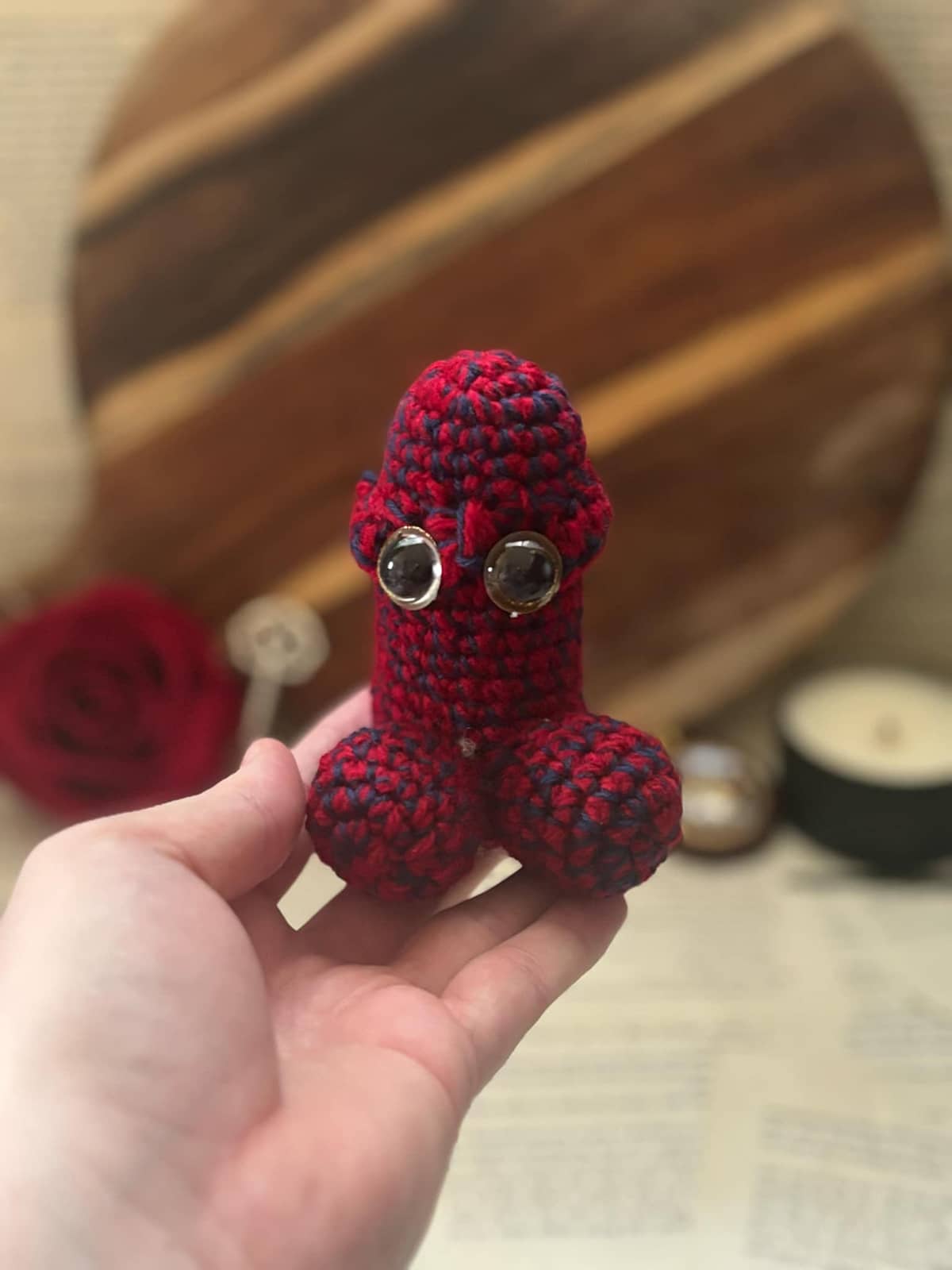 Hand holding crochet pin cushion in penis shape, featuring eyes on bright red and black yarn