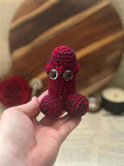 Hand holding crochet pin cushion in penis shape, featuring eyes on bright red and black yarn