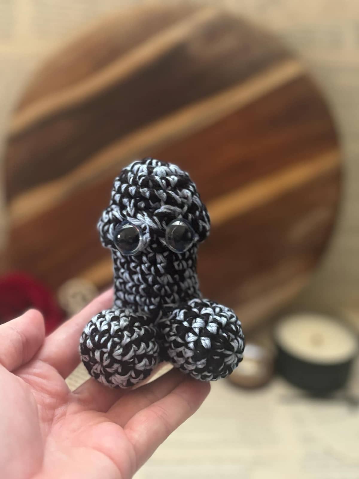 Hand holding crochet pin cushion in penis shape, featuring eyes on silver and black yarn