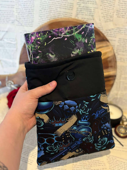 Hand holding standard book sleeve featuring blue and gold guns and roses design on black fabric, containing paperback book