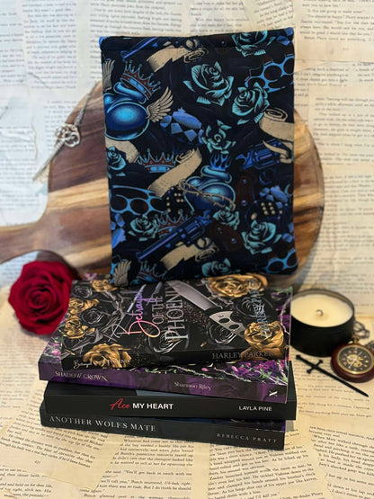 Standard book sleeve featuring blue and gold guns and roses design on black fabric, displayed on stack of books against scattered book pages