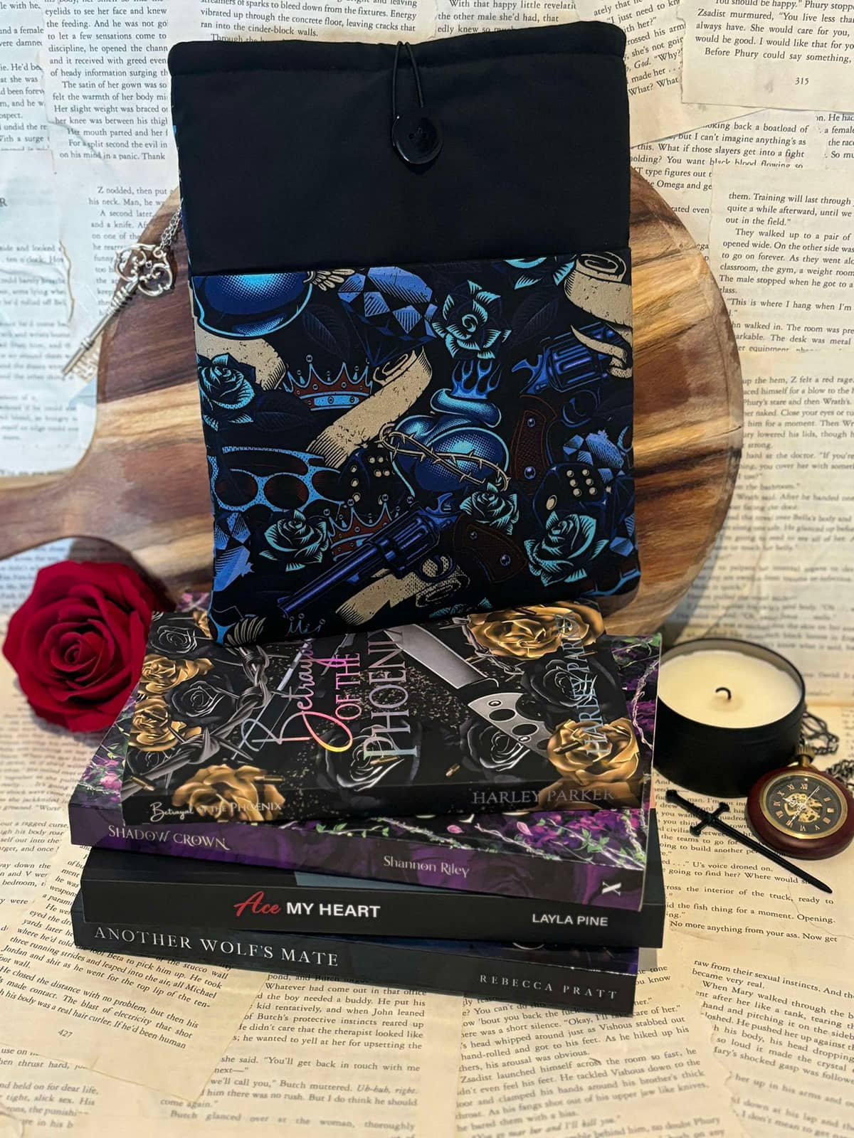 Standard book sleeve featuring blue and gold guns and roses design on black fabric, displayed on stack of books against scattered book pages