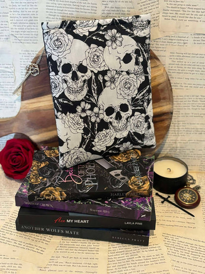 Standard book sleeve featuring white skulls and flowers design on black fabric, displayed on stack of books against scattered book pages