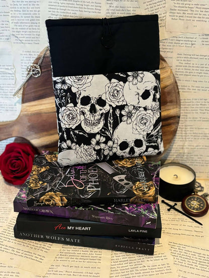 Standard book sleeve featuring white skulls and flowers design on black fabric, displayed on stack of books against scattered book pages