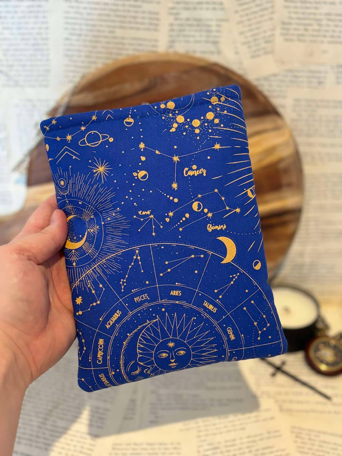 Hand holding Kindle sleeve featuring gold astrology design on cobalt blue fabric, displayed against scattered book pages