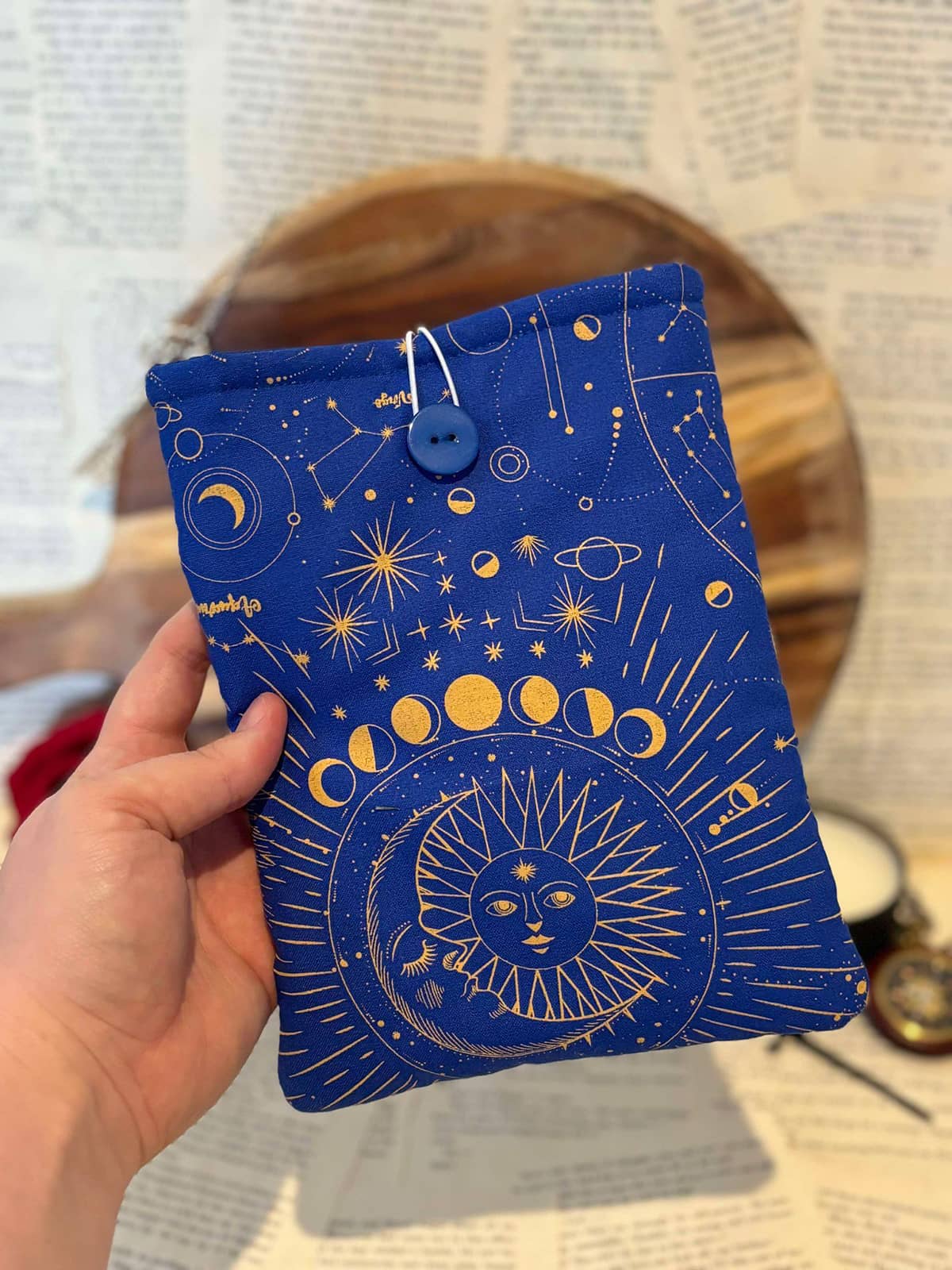 Hand holding Kindle sleeve featuring gold astrology design on cobalt blue fabric, displayed against scattered book pages