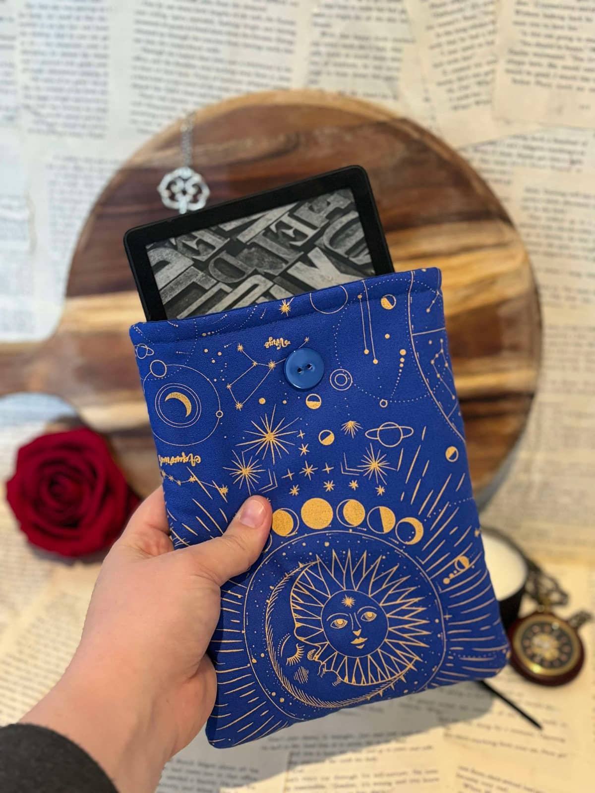 Hand holding Kindle sleeve featuring gold astrology design on cobalt blue fabric, containing Kindle device inside