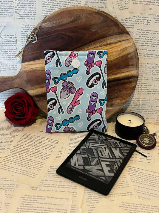 Kindle sleeve featuring light blue fabric with female adult toys design, displayed against scattered book pages next to a Kindle device