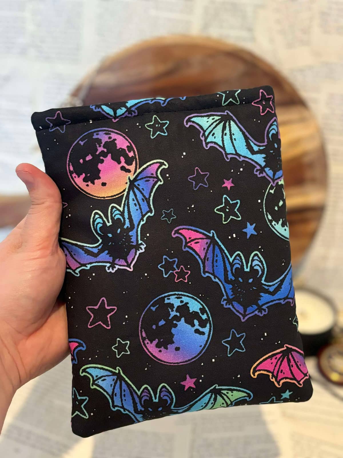 Hand holding Kindle sleeve featuring neon bats and stars design on black fabric, displayed against scattered book pages