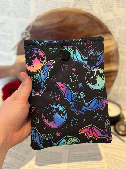 Hand holding Kindle sleeve featuring neon bats and stars design on black fabric, displayed against scattered book pages