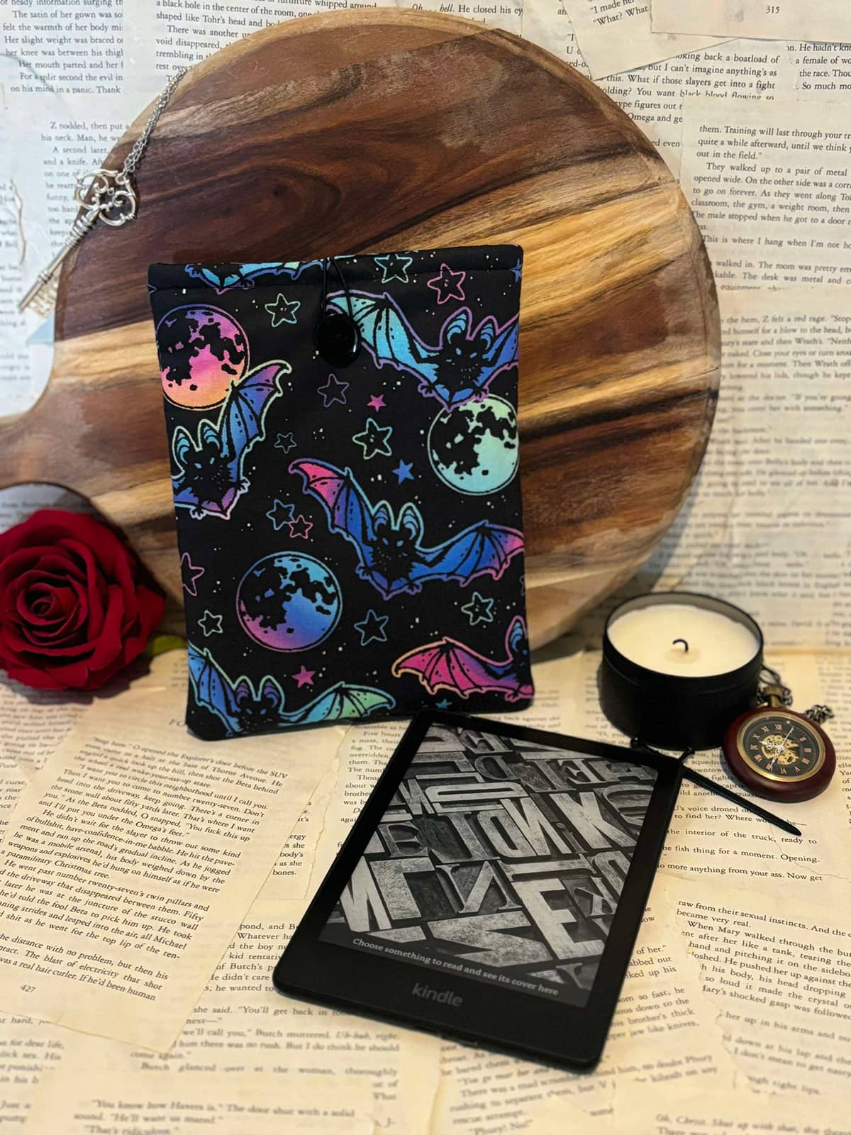 Hand holding Kindle sleeve featuring neon bats and stars design on black fabric, displayed against scattered book pages next to Kindle device