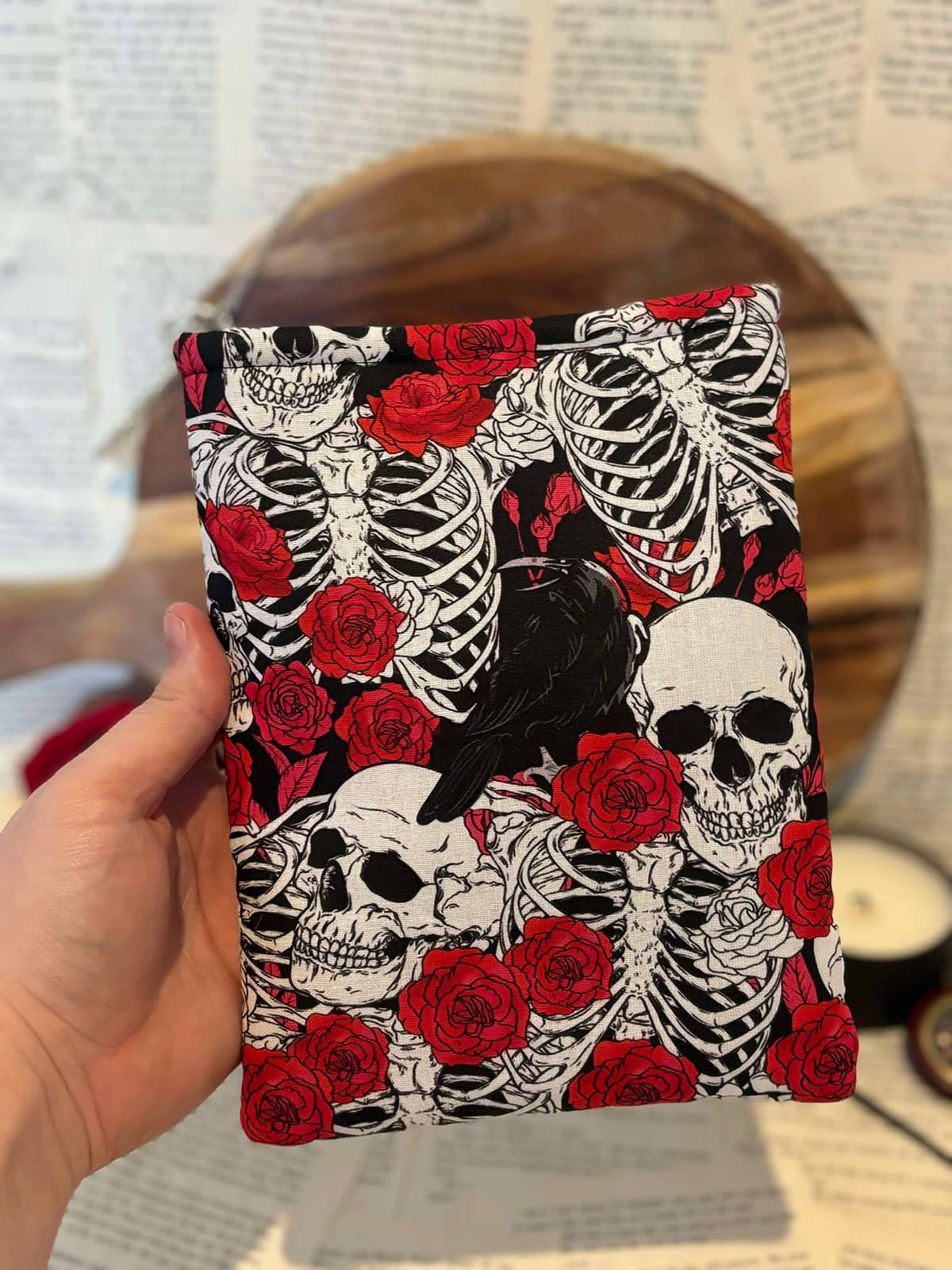 Hand holding Kindle sleeve featuring red roses and white skulls design on black fabric, displayed against scattered book pages