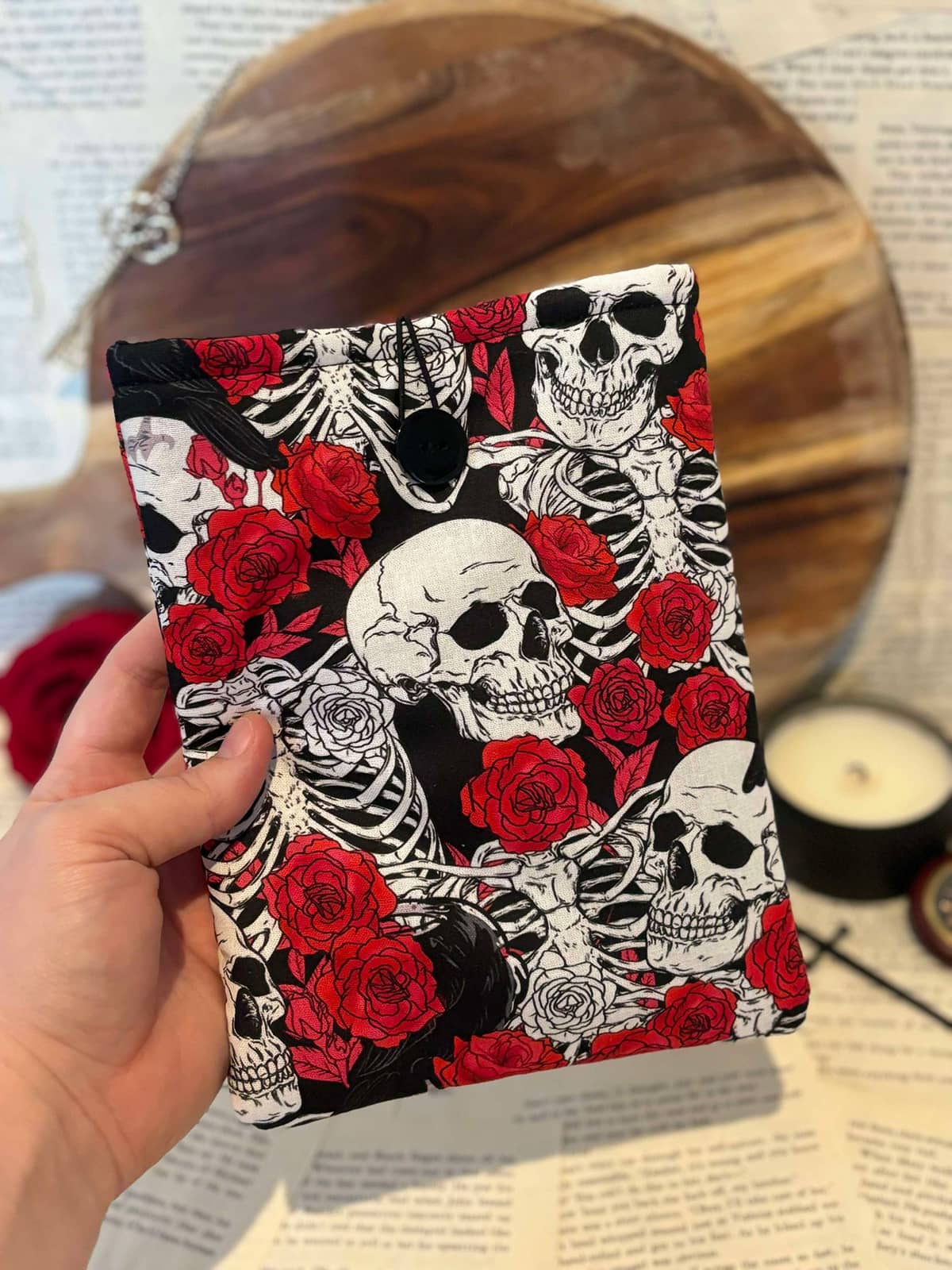 Hand holding Kindle sleeve featuring red roses and white skulls design on black fabric, displayed against scattered book pages