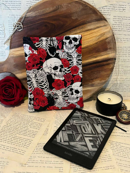 Kindle sleeve featuring red roses and white skulls design on black fabric, displayed against scattered book pages next to Kindle device