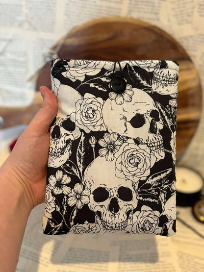 Hand Holding Kindle sleeve featuring white skulls and flowers design on black fabric, displayed against scattered book pages 