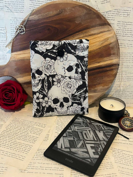 Kindle sleeve featuring white skulls and flowers design on black fabric, displayed against scattered book pages next to Kindle device