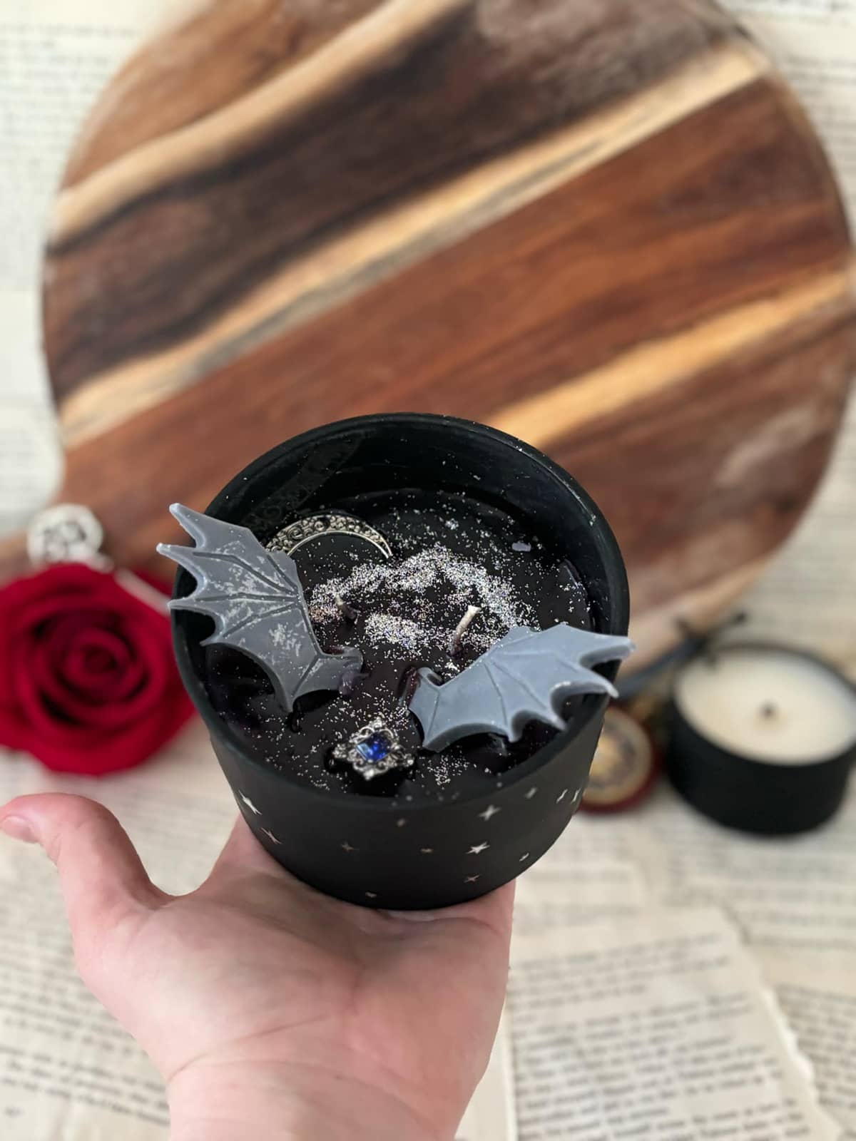 Hand holding candle featuring black wax with silver glitter, silver moon, silver charm with blue gem, and grey wings, set in a matte black vessel with starry design