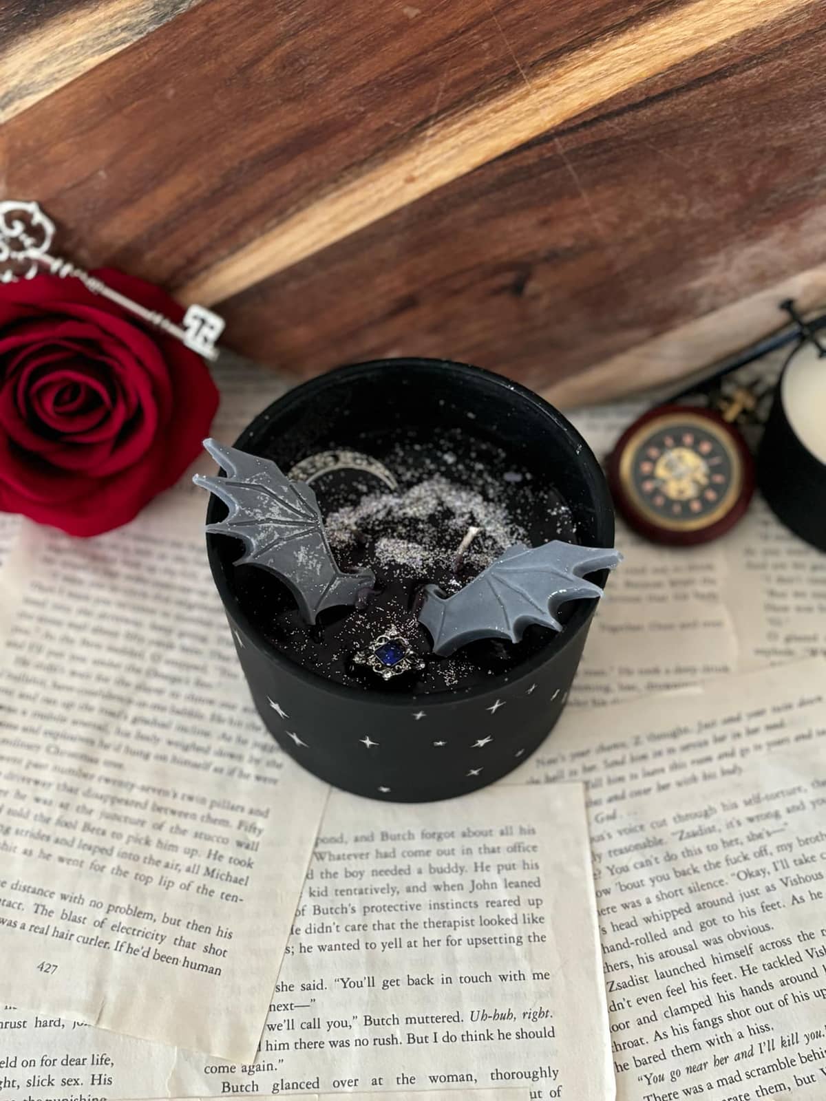 Candle featuring black wax with silver glitter, silver moon, silver charm with blue gem, and grey wings, set in a matte black vessel with starry design