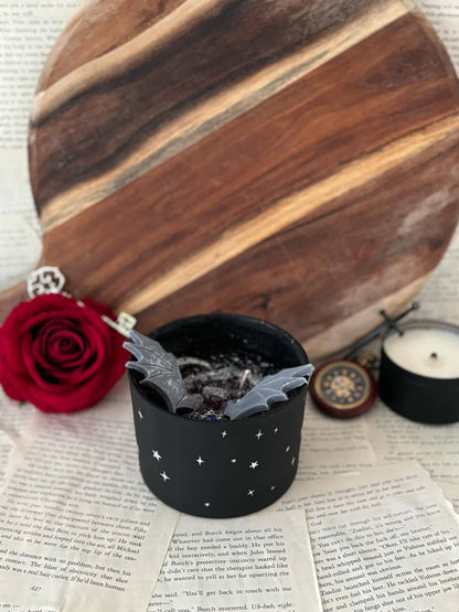 Candle featuring black wax with silver glitter, silver moon, silver charm with blue gem, and grey wings, set in a matte black vessel with starry design